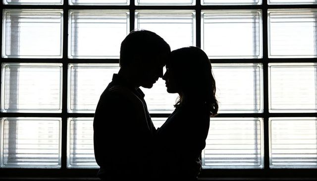 Silhouetted couple embracing before glass block window, intimate backlit romance