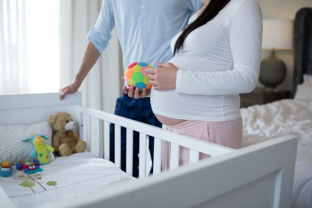 Expecting parents standing near a baby crib, holding colorful toys, and preparing the nursery for their upcoming baby. This image can be used in articles or advertisements related to pregnancy, parenthood, baby products, family bonding, and home preparation for a newborn.