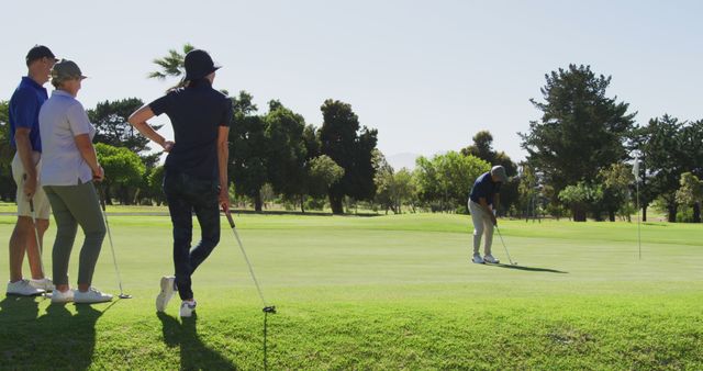 Group of golfers observes team member putting on pristine green golf course under sunny sky. Perfect for use in articles about outdoor sports, recreational activities, group outings, teamwork, and golf training. Highlights the serene environment and interactive nature of the sport.