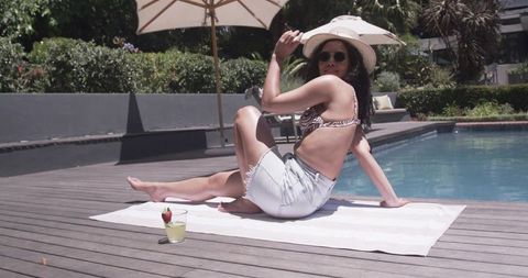 African woman relaxing by pool wearing sunhat and denim cutoffs holding cocktail luxury resort