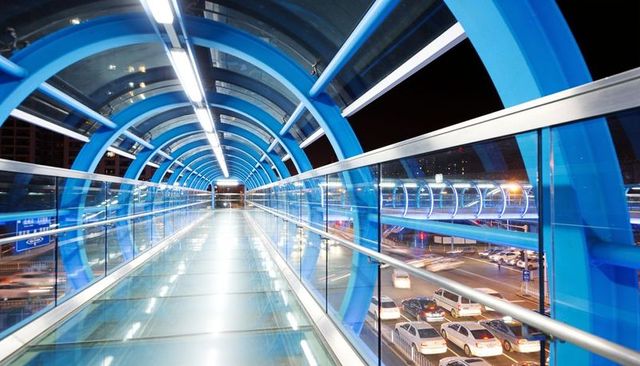 Blue-lit enclosed pedestrian bridge with glass floor and arched tunnel over night traffic