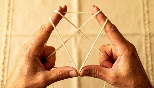 Adult hands weaving cat's cradle with white twine on beige woven surface closeup