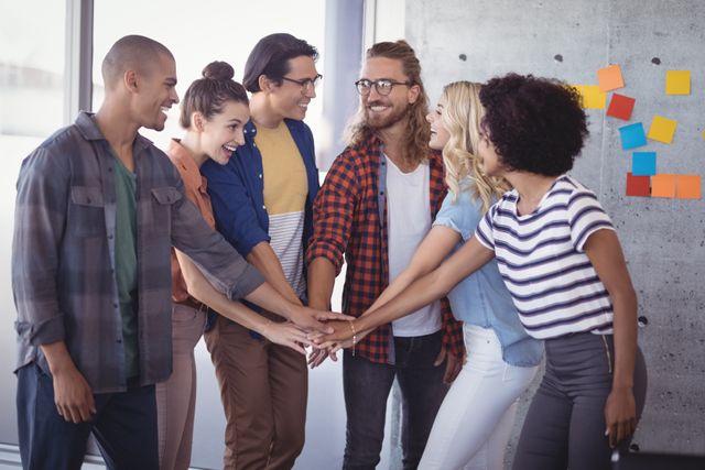 Diverse group of young professionals stacking hands in a creative office environment, symbolizing teamwork, unity, and collaboration. Ideal for use in articles or advertisements related to business success, team building, startup culture, and motivational content.