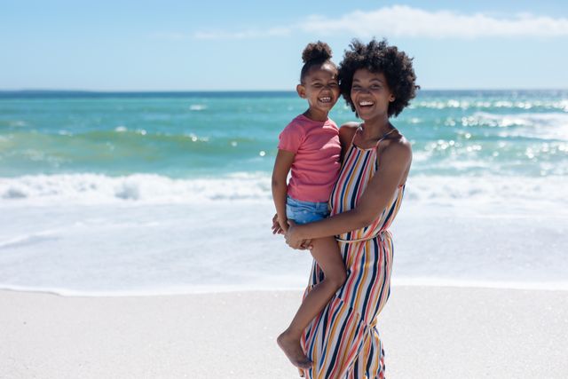Portrait of smiling african american mother carrying daughter standing at beach on sunny day. unaltered, family, lifestyle, togetherness, enjoyment and holiday concept.