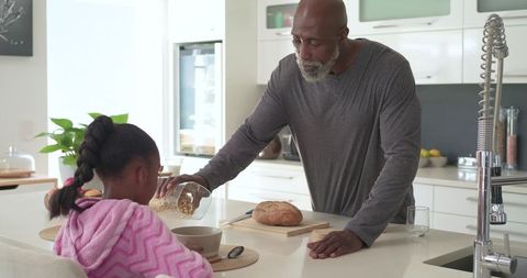 Senior african american man pouring cereal for daughter at modern kitchen island morning routine