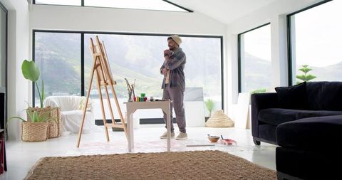 Indian artist standing and contemplating canvas in sunlit modern living room studio