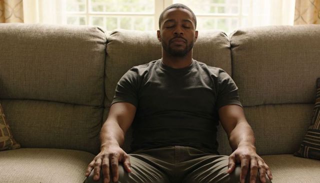 Man meditating on sofa with eyes closed, daytime home relaxation and calm mindfulness