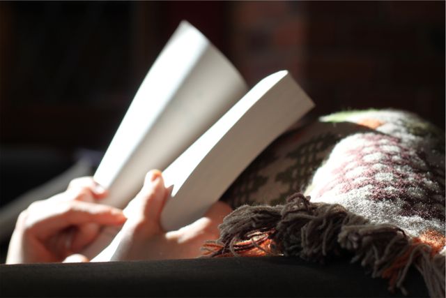 Hands holding book, covered with patterned blanket, sunlight streaming in, creating a warm and cozy atmosphere, great for themes of relaxation, home comfort, leisure activities, and peaceful afternoons. Useful for blogs, lifestyle websites, mental health content, book clubs, and social media posts promoting self-care and calmness.
