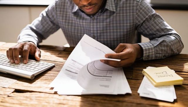 Man reviewing invoices and bills at wooden desk with keyboard, sticky notes, paperwork