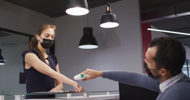 Caucasian woman in face mask has temperature checked by male colleague at office reception desk. independent business at a modern office during coronavirus covid 19 pandemic.
