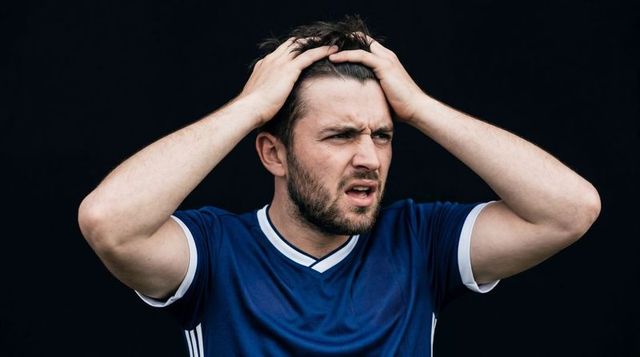 Frustrated bearded athlete holding head and reacting in blue jersey on dark background