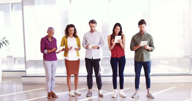 Group of young multiracial professionals standing in modern office, using tablets and phones. Ideal for illustrating concepts such as modern workplace, teamwork, communication technology, and multicultural office environments. Suitable for business presentations, team-building seminars, and articles about workplace diversity and innovation.
