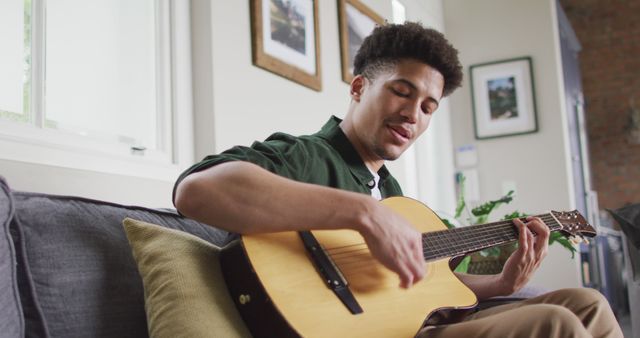 Young man sitting on couch playing acoustic guitar in modern living room. Perfect for use in advertisements, articles or websites focused on music, relaxation, creativity, or modern home living concepts.