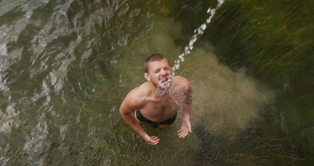 Young man standing under a refreshing spray from waterfall in forest stream. Perfect for advertisements promoting adventure travel, outdoor activities, vacations, natural beauty, or lifestyle blogs focusing on wellness and nature.