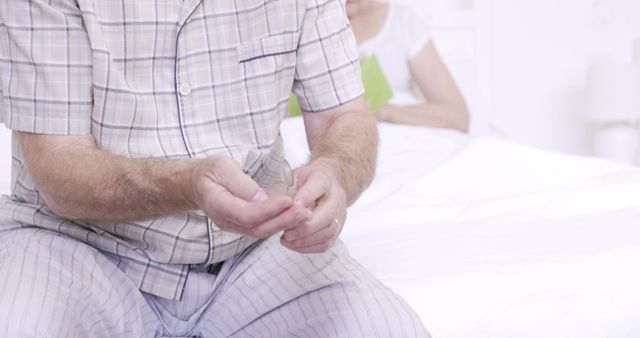 Elderly man is sitting on edge of bed, dressed in comfortable pajamas. He is holding his hands and appears to be contemplating. This scene is in a bedroom with another person blurred in the background on the bed. This photo is useful for topics related to senior health, caregiving, morning routines, and elderly care products.
