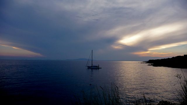 A single sailboat resting on calm waters as the sun sets and casts an orange glow with hints of purples and blues over the horizon. Ideal for travel blogs, adventure imagery, inspirational and motivational materials, and serene landscape collections.