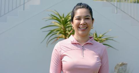 Smiling woman wearing pink quarter-zip top standing in sunlit courtyard with greenery