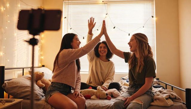 High-fiving friends on bed during cozy bedroom hangout with phone tripod and string lights