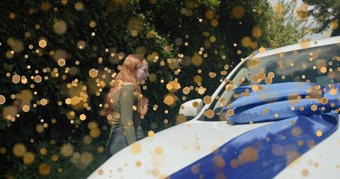 Redheaded woman standing beside white suv wrapped in blue ribbon with golden bokeh