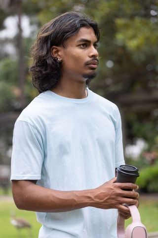 Young man standing in park holding takeaway coffee and pink headphones outdoor portrait