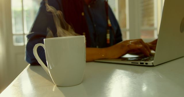 Close-up of steam coming out of coffee cup on dining table in kitchen of comfortable home. Black woman working on laptop 4k