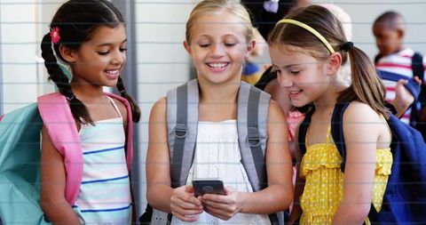 Smiling schoolgirls gathering around smartphone in school corridor with backpacks