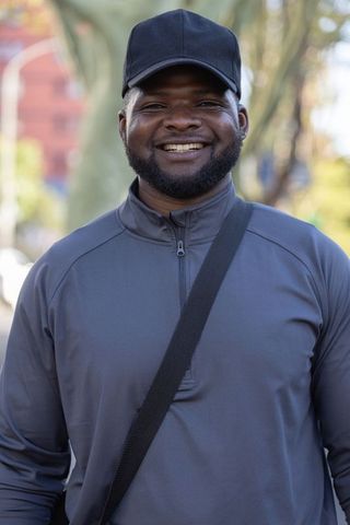 African man smiling while walking on tree-lined urban street wearing cap and crossbody bag