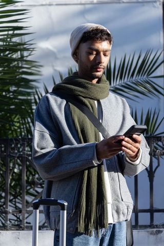 Man checking smartphone while traveling with suitcase and olive scarf in sunlit palm
