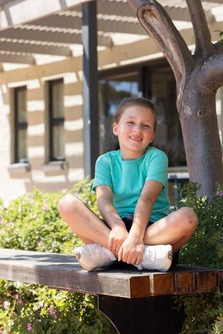 Smiling girl sitting cross-legged on wooden bench in sunlit courtyard wearing teal shirt