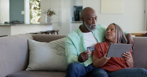Mature diverse couple shopping online together using tablet and credit card on couch