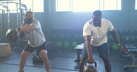 Two african men swinging kettlebells during partner workout in bright industrial gym