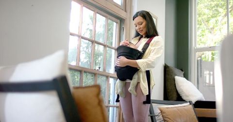 Asian mother nurturing infant in carrier by sunlit window