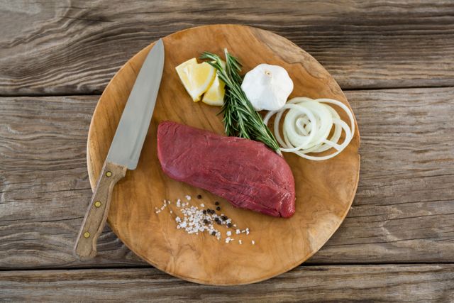 Raw beef steak placed on a wooden tray with fresh ingredients including garlic, rosemary, lemon, onion, salt, and pepper. A knife is also present, suggesting preparation for cooking. Ideal for use in culinary blogs, recipe websites, cooking tutorials, and food-related advertisements.