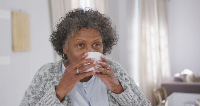 Elderly African American woman is drinking a hot beverage while at home, wearing a cozy sweater and pajama top. Image can be used for health and wellness articles, self-care promotions, or senior living brochures.