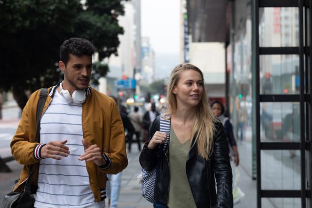 Happy, fashionable Caucasian couple out and about walking in the city street during the day, talking and smiling, man wearing wireless headphones around his neck.