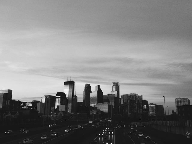 A monochromatic view of a downtown cityscape at dusk. The high-rise buildings and office structures create a dramatic skyline. Cars and traffic are visible on the road in the foreground. This image can be used for urban planning presentations, city guides, travel brochures, architectural designs, and real estate advertisements.