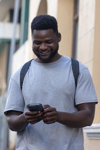 Smiling man checking smartphone with backpack on city sidewalk in gray tee