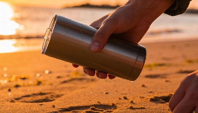 Holding stainless steel tumbler on sandy beach at sunset with water droplets and glare