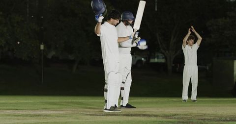 Night cricket players raising helmet and bat celebrating milestone on floodlit pitch