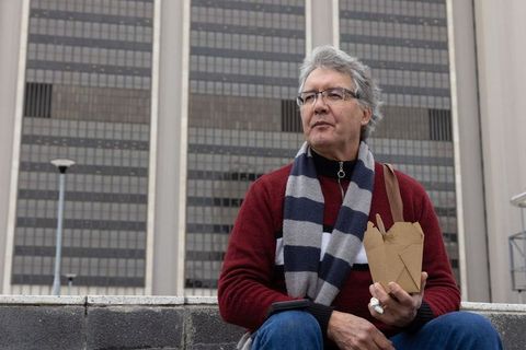 Senior man eating takeout lunch on urban plaza ledge wearing scarf and glasses, office tower
