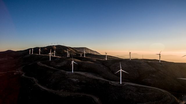 Wind turbines are scattered across a hilly landscape during sunset, highlighting the importance of renewable energy. Perfect for illustrating themes of sustainable development, clean energy solutions, and environmental conservation. Suitable for use in articles about renewable energy, educational materials, environmental campaigns, and presentations on sustainable practices.