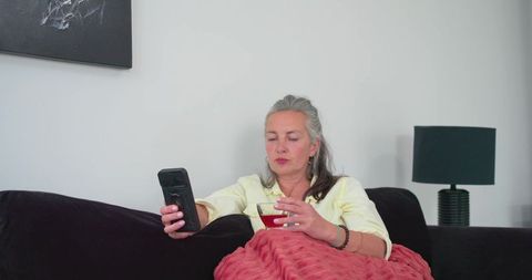 Mature woman relaxing on couch with smartphone and wine glass in modern living room
