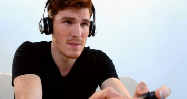 A young man with headphones is gaming on a console. His expression shows concentration and engagement. This image can be used for content related to gaming, technology, or electronic sports. It is also suitable for illustrating the lifestyle of gamers and entertainment at home.