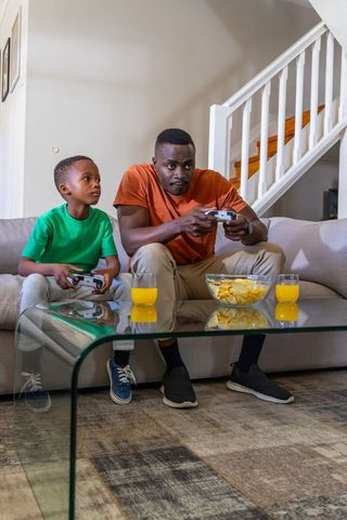 African american father and son playing video games on couch with chips and juice