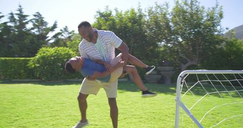 African american father spinning son on backyard lawn near soccer goal family fun