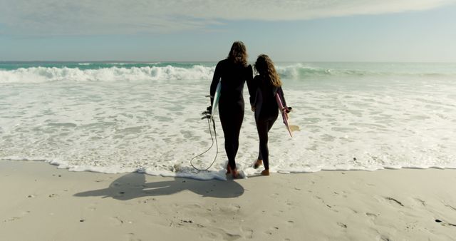 Mother and daughter walking on beach with their surfboards, wearing wetsuits, embraced while heading towards ocean waves. Perfect for promoting family vacations, water sports, surf training, unity, and outdoor activities. Shows warmth and strong family bonds.