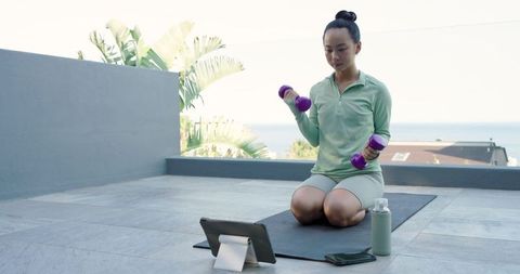 Woman Exercising with Dumbbells on Patio Watching Tablet