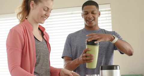 Young couple making green smoothie in bright home kitchen with personal blender cup