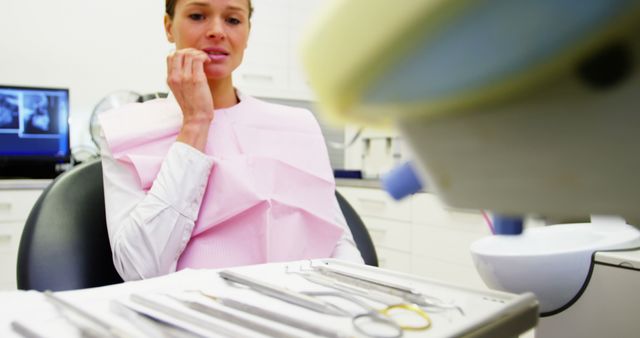 Patient sitting in the dentist's office with nervous expression, anticipating dental examination. Dental tools visible in foreground, adding to the clinical atmosphere. Suitable for use in healthcare articles, dental care advertisements, anxiety and mental health articles, blogs about dental experiences, and medical publications.