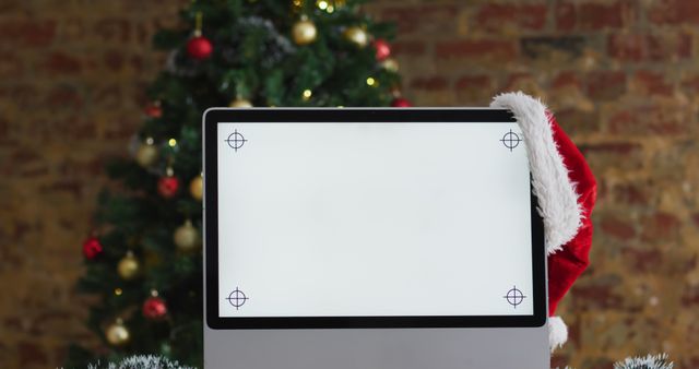 Blank computer screen adorned with a Santa hat positioned in front of a Christmas tree. Perfect for holiday-themed corporate greetings, festive workspace promotions, and seasonal technology marketing materials.